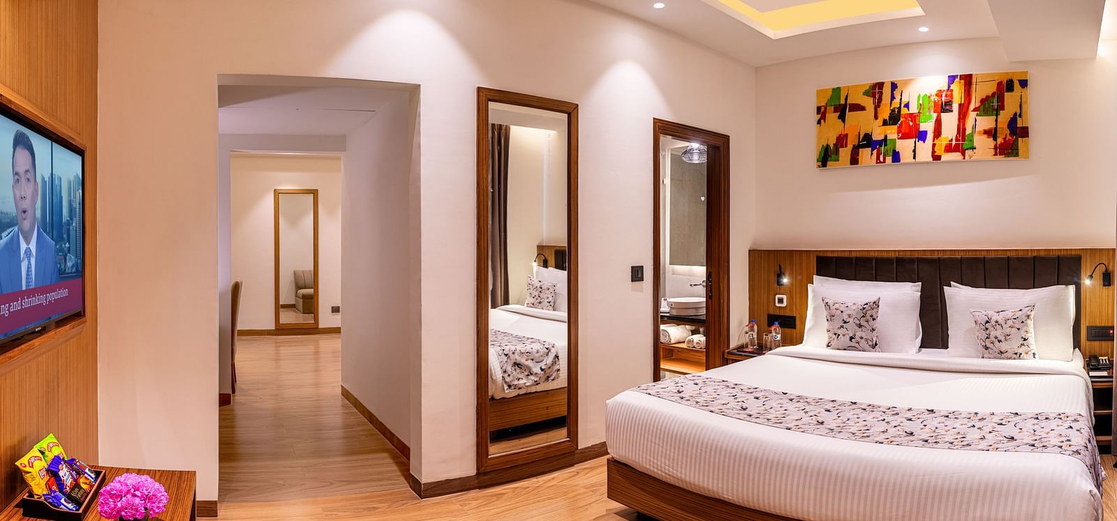 Hotel room with a king size bed, flat-screen TV, and a hallway leading to another room. It features a unique tray ceiling with lighting, a ceiling fan, and colourful abstract artwork - Lords Eco Inn Dehradun