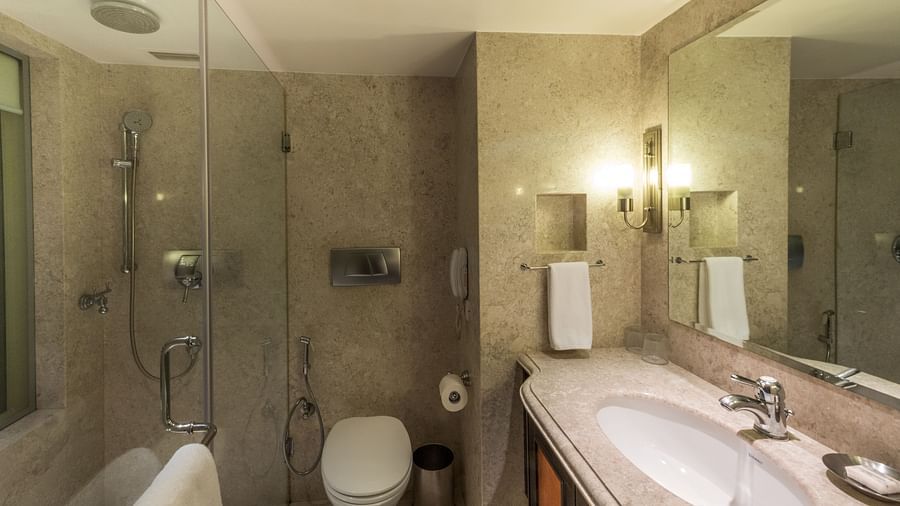 Modern hotel bathroom with a walk-in shower, toilet, twin sinks, and well-lit mirrors.