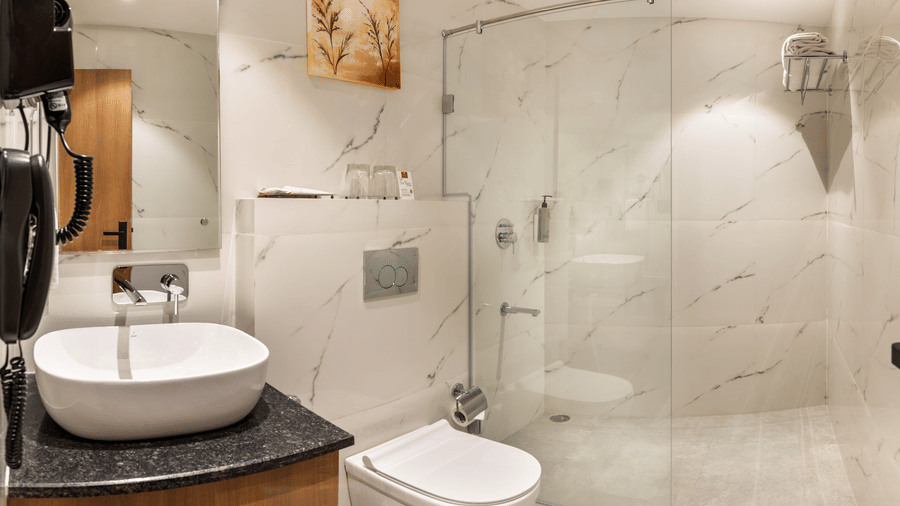 A contemporary bathroom at Lords Inn Katra featuring a white washbasin, a wall mounted toilet, a mirror, and a glass shower cubicle.
