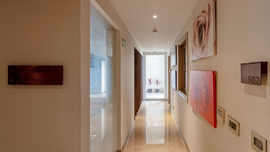A modern corridor lined with elegant paintings on the right wall, leading to a glass door at the end that allows light to illuminate the hallway.