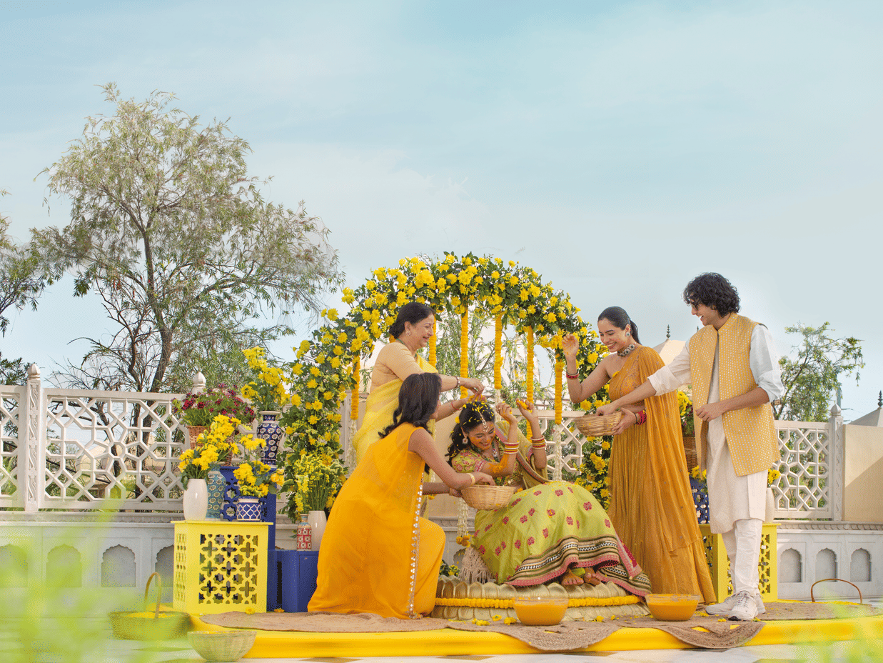 A joyful outdoor family gathering with four individuals around a decorative floral arch, engaging happily in haldi ceremony on a sunny day at Heritage Village Resorts & Spa, Manesar - a Resort Near Delhi for Wedding.