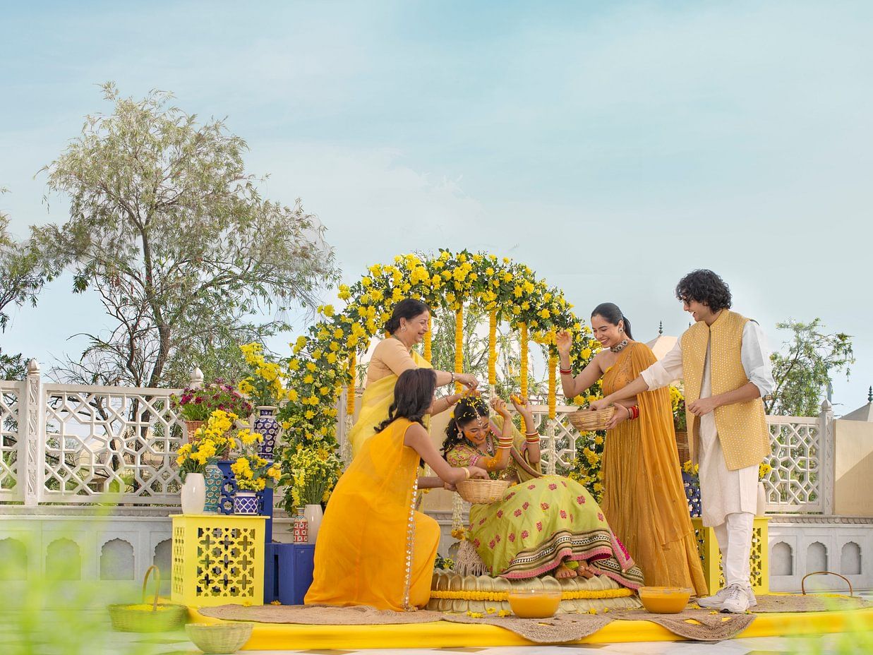 A joyful outdoor family gathering with four individuals around a decorative floral arch, engaging happily in haldi ceremony on a sunny day.