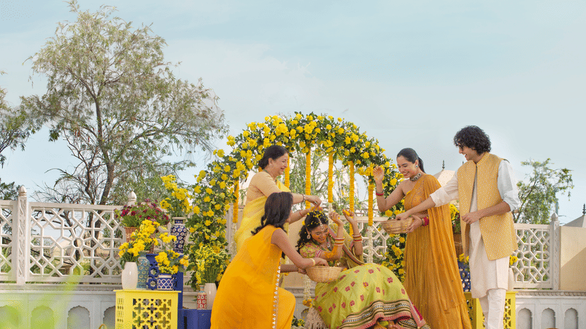 A joyful outdoor family gathering with four individuals around a decorative floral arch, engaging happily in haldi ceremony on a sunny day at Heritage Village Resorts & Spa, Manesar - a Resort Near Delhi for Wedding.