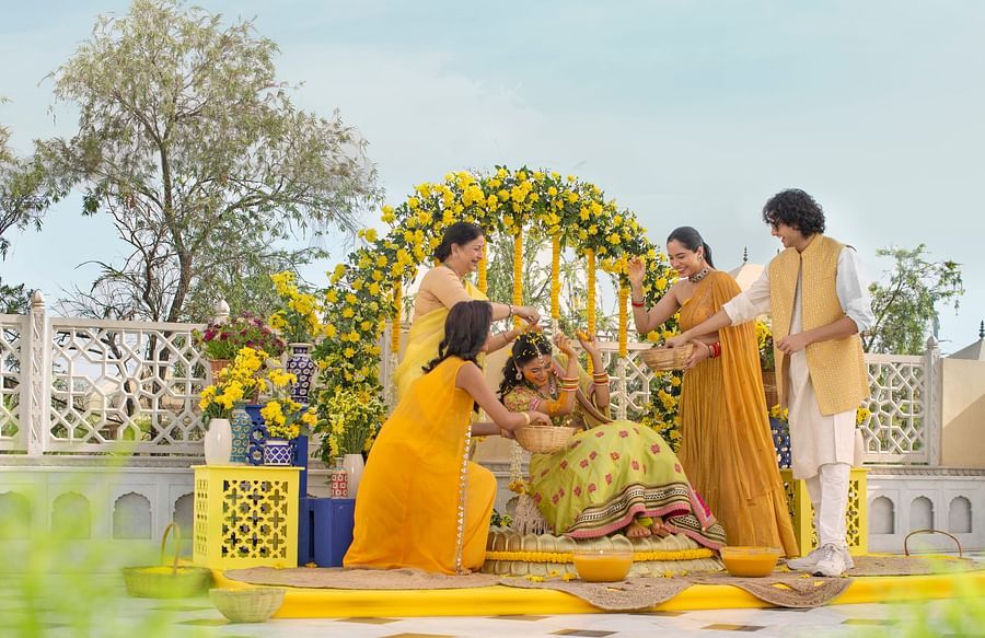 A joyful outdoor family gathering with four individuals around a decorative floral arch, engaging happily in haldi ceremony on a sunny day.