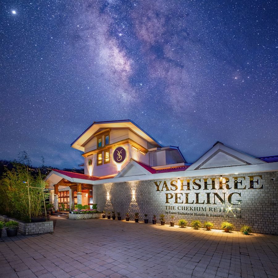 Evening view of Yashshree Pelling hotel, featuring a brightly lit entrance, stone façade, and red-roofed building under a deep blue sky.
