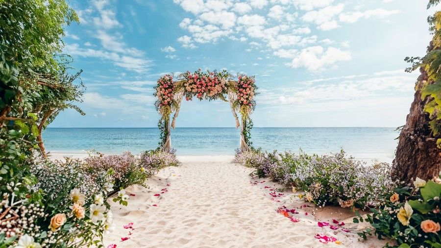 Wedding area near beachside featuring a sandy pathway and on either side there are decorations of flowers and leaves. At the end of the pathway there is a wooden frame acting as a stage decorated with flowers.