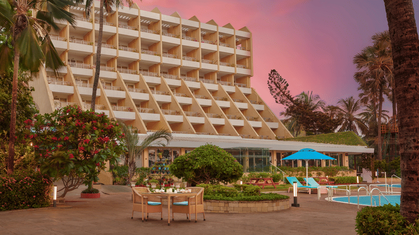 An exterior view of The Resort, Mumbai, with palm trees, a pool, and a gradient sky in the background.
