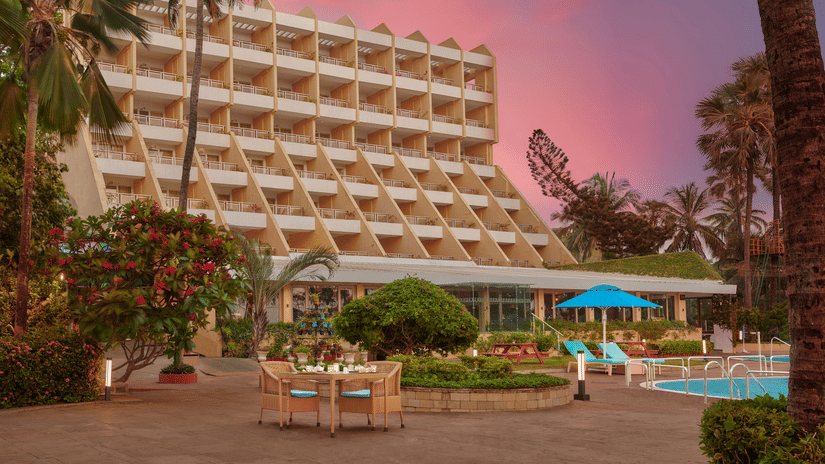 An exterior view of The Resort, Mumbai, with palm trees, a pool, and a gradient sky in the background.