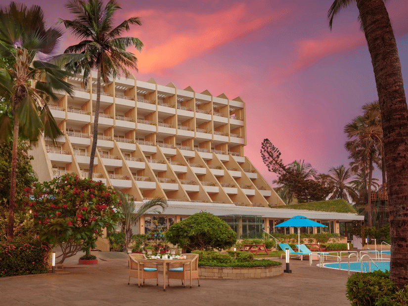An exterior view of The Resort, Mumbai, with palm trees, a pool, and a gradient sky in the background.
