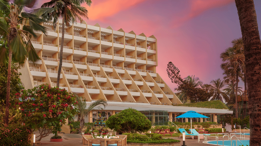 An exterior view of The Resort, Mumbai, with palm trees, a pool, and a gradient sky in the background.