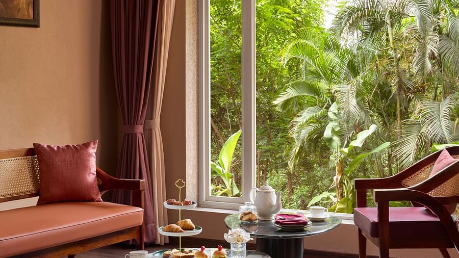 The living room of Junior Suite at DGV Resort by Treat, Silvassa with a comfortable sofa, a coffee table with snacks, and a large window overlooking lush greenery