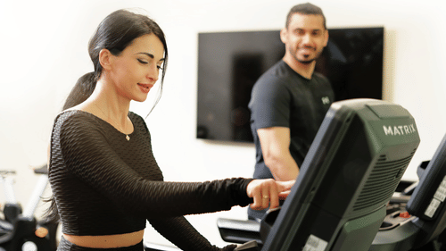 a couple inside the gymnasium on a treadmill while the man looks at the woman