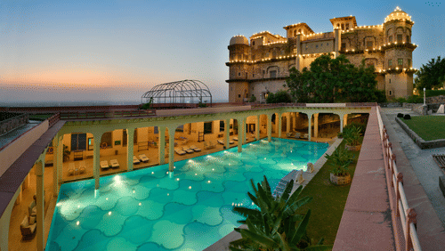 An aerial view of a swimming pool at dusk, with the historic fort-palace illuminated in the background, showcasing Tijara Fort-Palace - 19th Century, Alwar