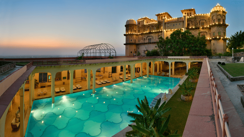 An aerial view of a swimming pool at dusk, with the historic fort-palace illuminated in the background, showcasing Tijara Fort-Palace - 19th Century, Alwar