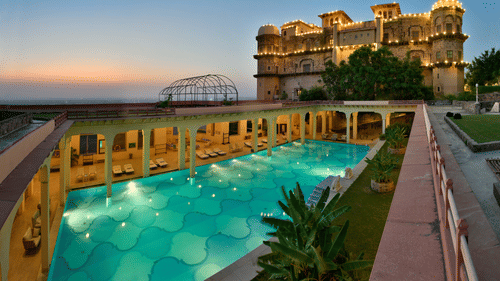 An aerial view of a swimming pool at dusk, with the historic fort-palace illuminated in the background, showcasing Tijara Fort-Palace - 19th Century, Alwar