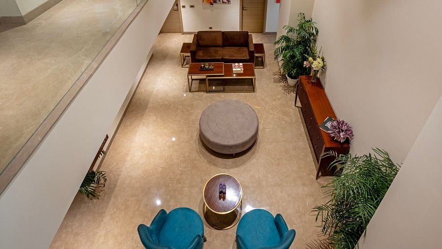 From above, the central seating in Hotel Saket 27’s lobby reveals a round table surrounded by two coffee tables, with a brown table against the wall. The corners are adorned with plants in small white pots, creating a balanced and inviting space.