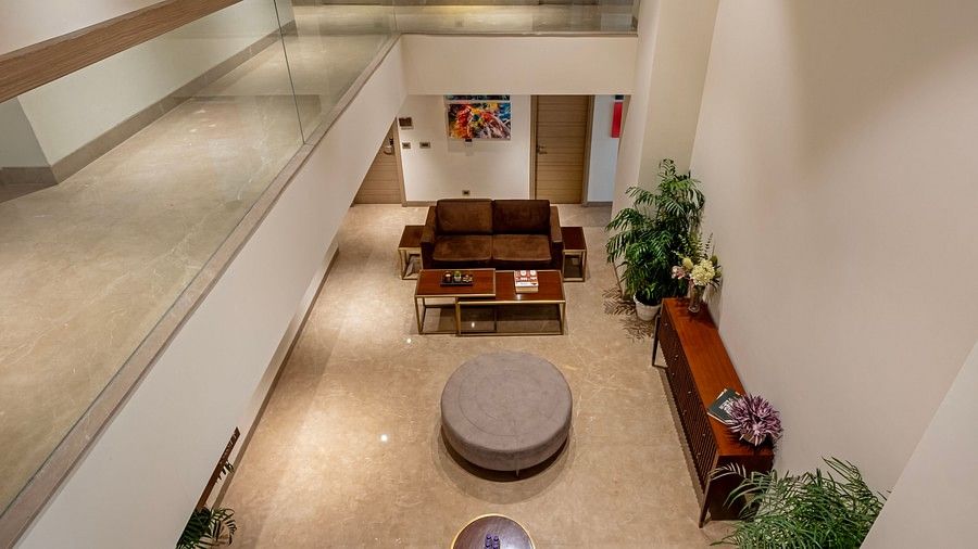 From above, the central seating in Hotel Saket 27’s lobby reveals a round table surrounded by two coffee tables, with a brown table against the wall. The corners are adorned with plants in small white pots, creating a balanced and inviting space.