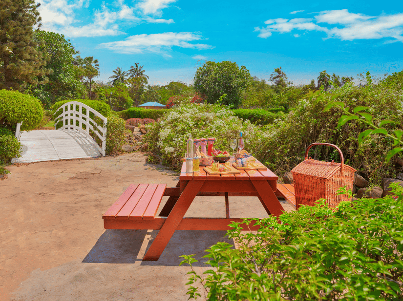 Outdoor garden picnic setup at The Resort, Mumbai with a wooden table, food spread, wicker basket, lush greenery, and a small white bridge under a bright blue sky.
