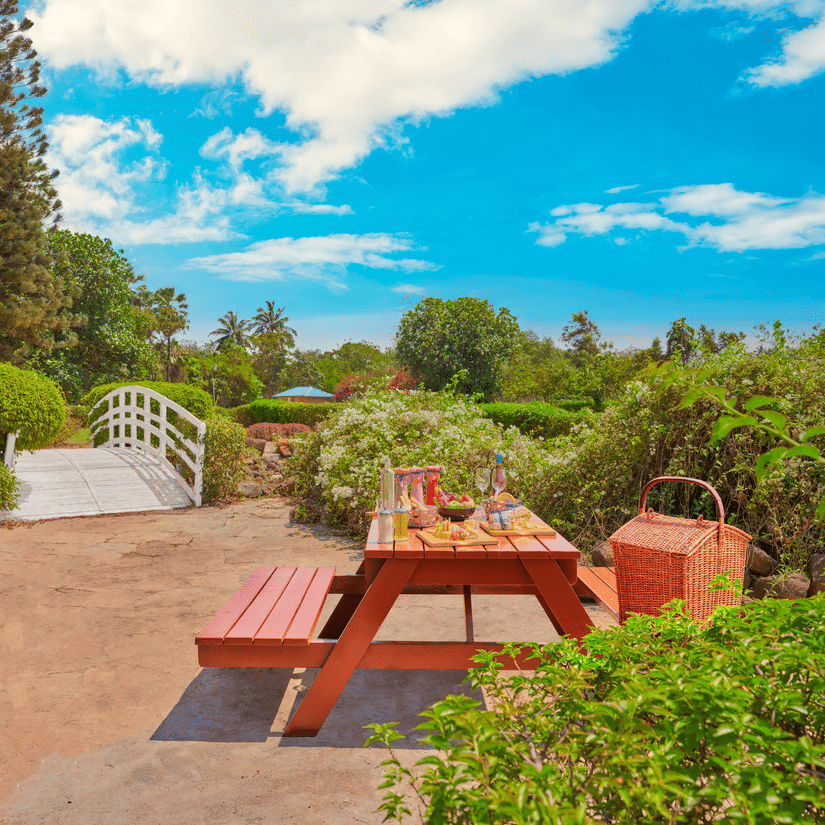 Outdoor garden picnic setup at The Resort, Mumbai with a wooden table, food spread, wicker basket, lush greenery, and a small white bridge under a bright blue sky.