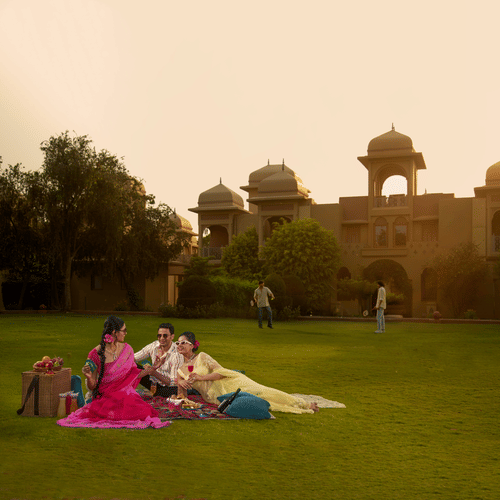 Three women in traditional indian attire are sitting in a lawn in front of a colorful, ornate building with an arched entrance. they appear to be engaged in a cheerful conversation.