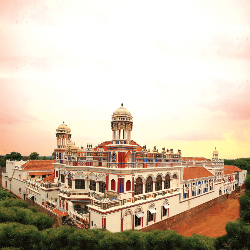 Facade view of Chidambara Vilas - A Luxury Chettinad Heritage Resort, surrounded by greenery and pink skies. 