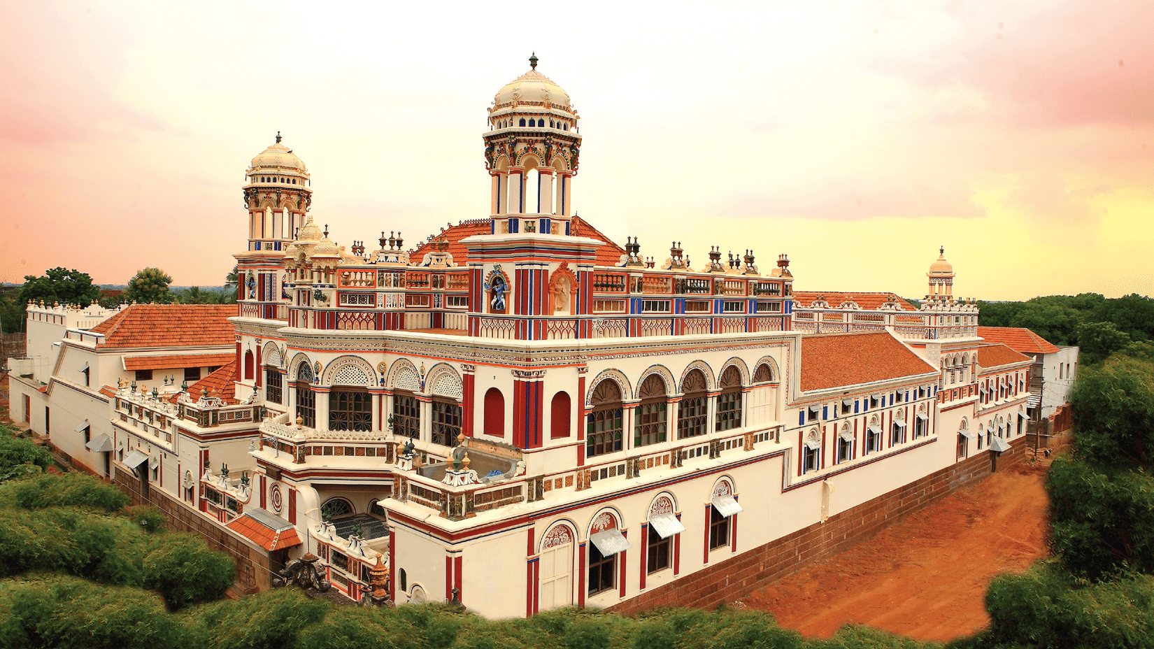 Facade view of Chidambara Vilas - A Luxury Chettinad Heritage Resort, surrounded by greenery and pink skies. 