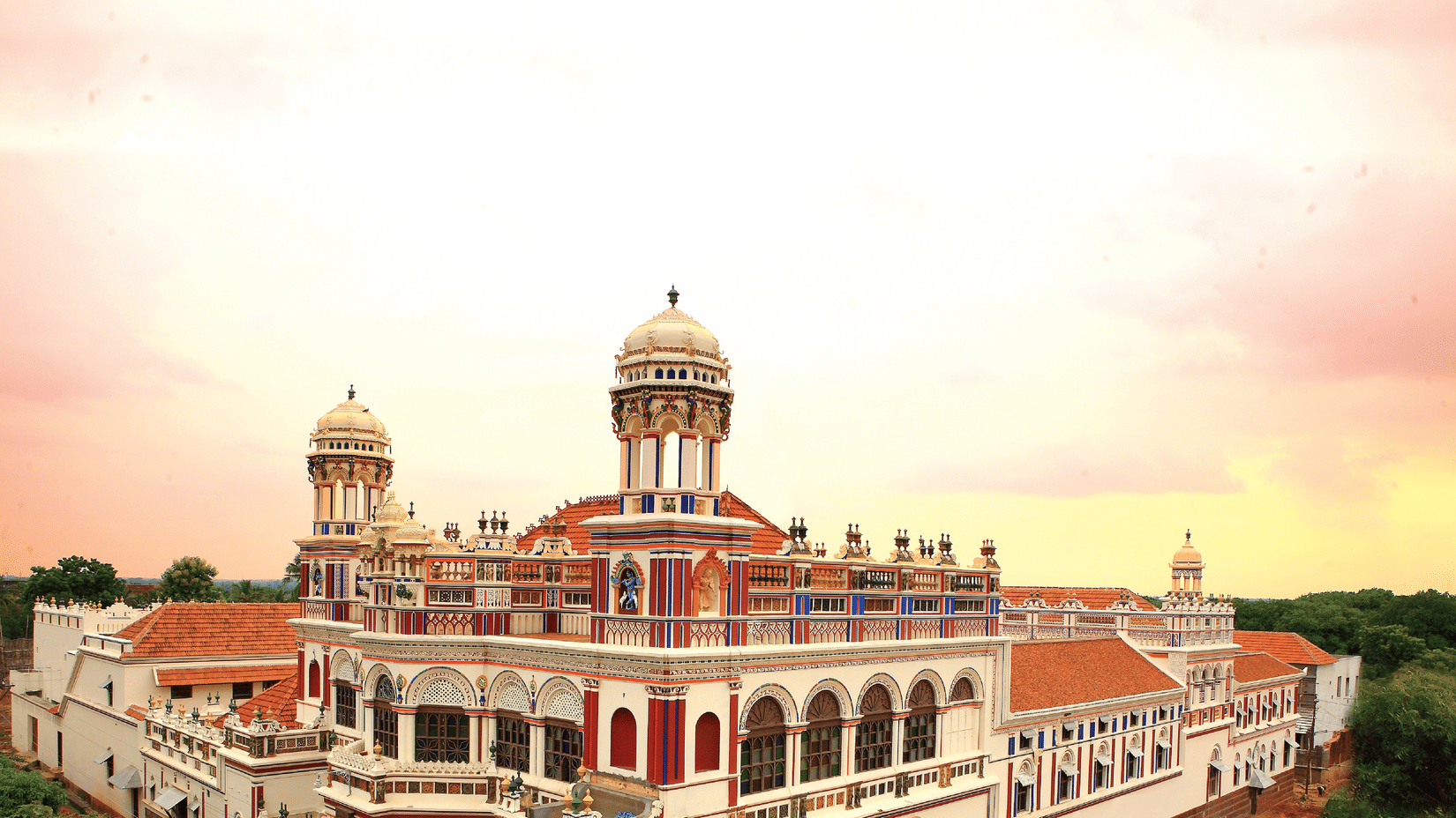 Facade view of Chidambara Vilas - A Luxury Chettinad Heritage Resort, surrounded by greenery and pink skies. 