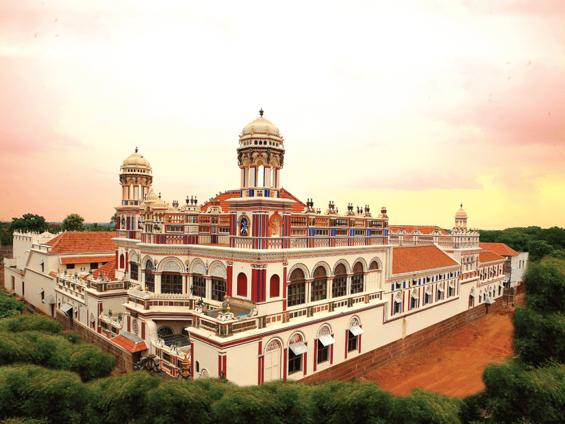 Facade view of Chidambara Vilas-Luxury Chettinad Heritage Resort, surrounded by greenery and pink sky. 