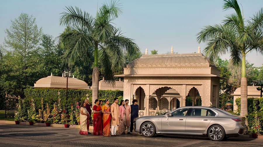 A traditional indian wedding scene outside a building with a couple dressed in bright orange and red attire standing beside a luxury car, surrounded by palm trees under a clear sky.