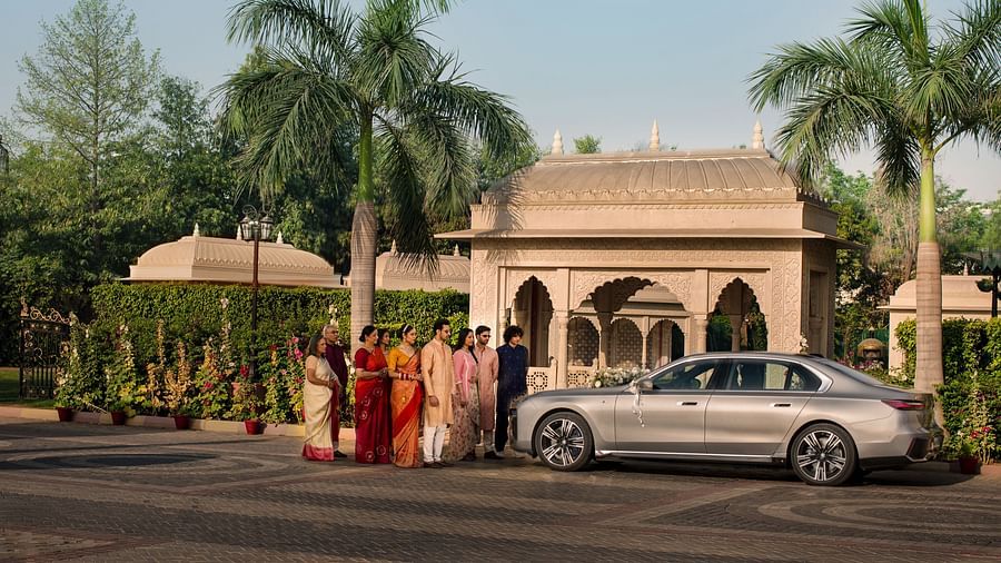 A traditional indian wedding scene outside a building with a couple dressed in bright orange and red attire standing beside a luxury car, surrounded by palm trees under a clear sky.