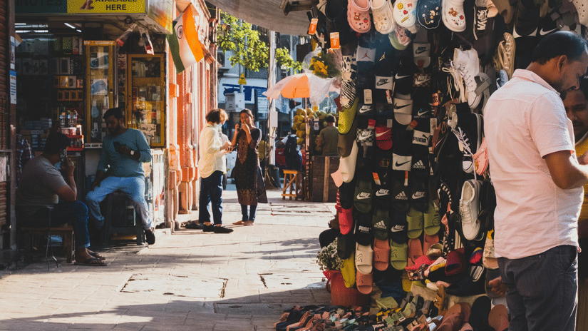 A busy narrow street market with a man in a white shirt browsing stalls selling shoes and accessories under a makeshift blue canopy.