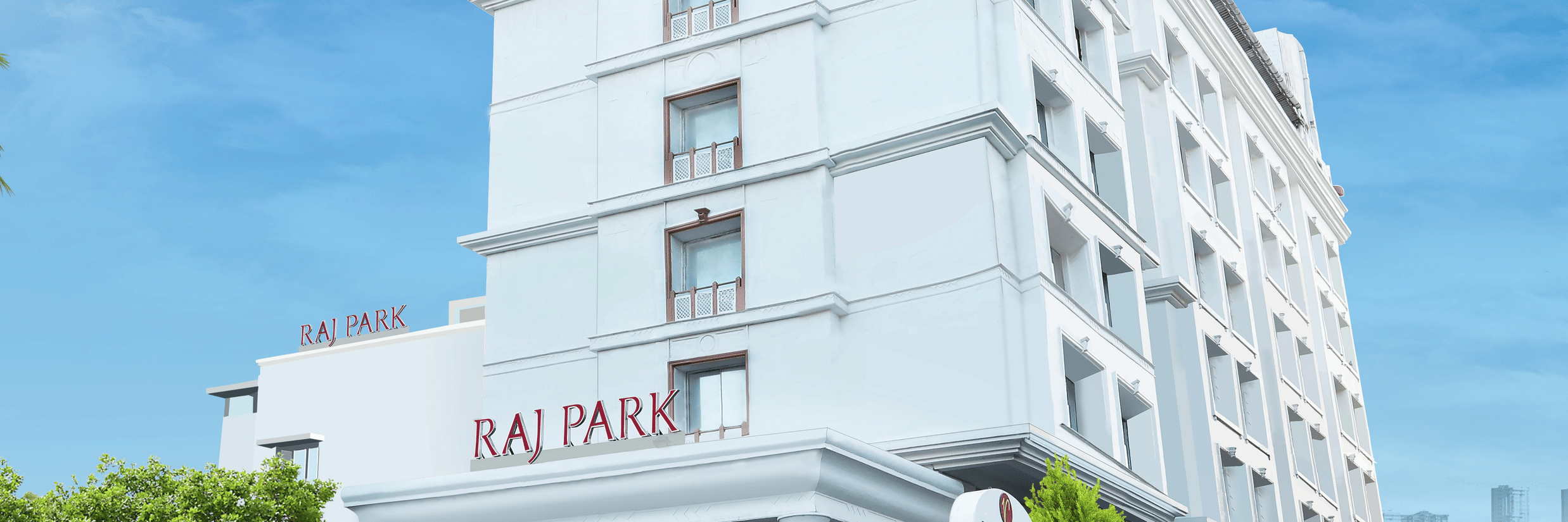 Facade of Raj Park hotel, one of the top hotels in Chennai, in the morning with blue sky and buildings in the background, surrounded by greenery.