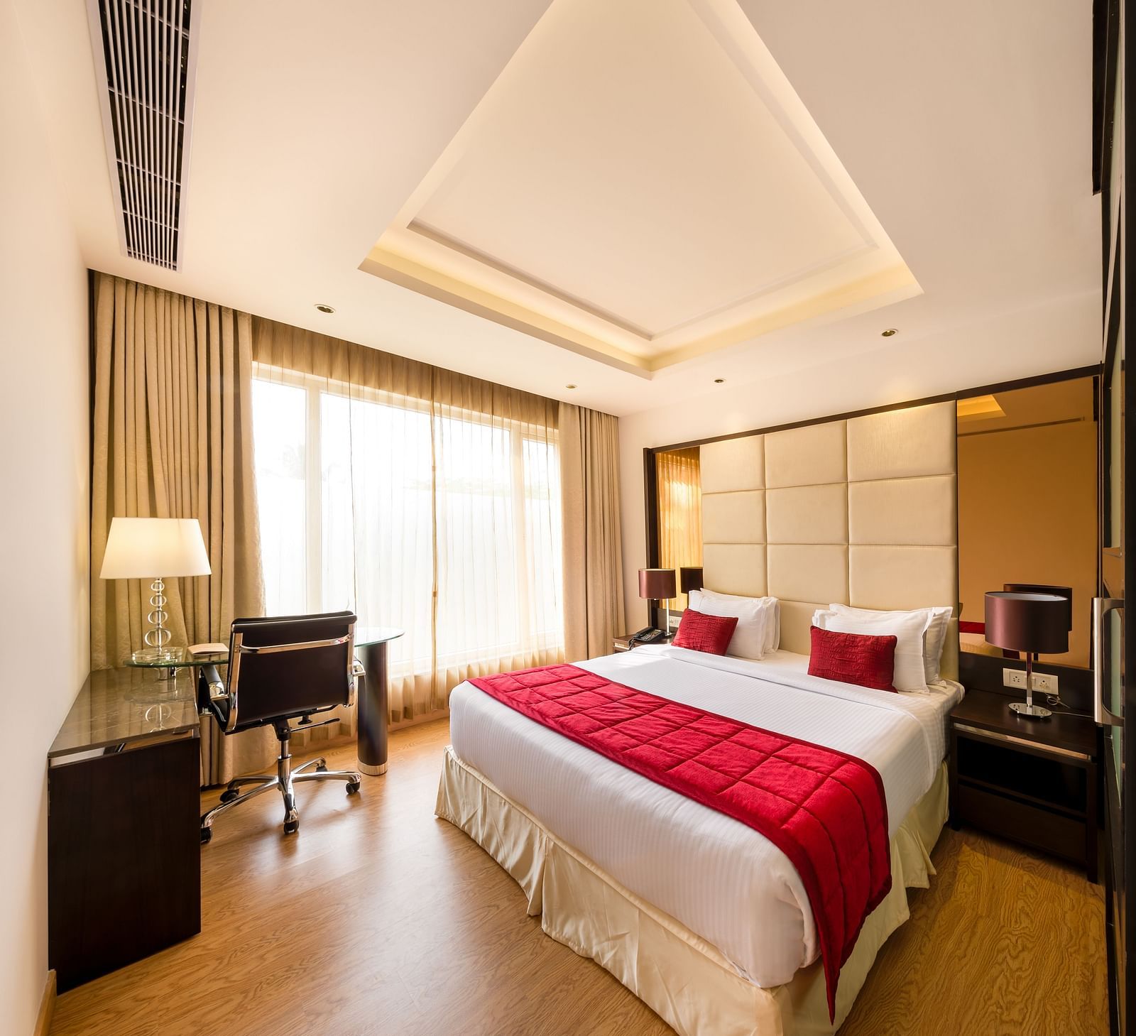 A look at the suite featuring a king size bed adorned with a red throw blanket, accompanied by a lamp and a desk with a chair - TGI Grand Fortuna, Hosur