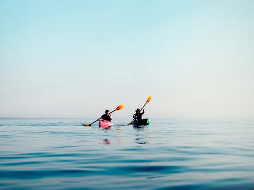Two kayakers paddling on calm blue water at sunrise