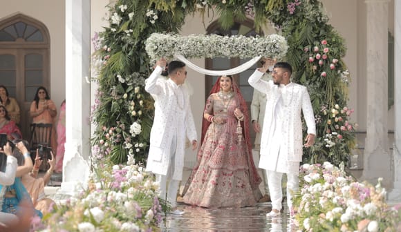 Bride walking down a mirrored aisle beneath a floral chadar held by two men in white sherwanis, framed by marble pillars at Fateh Vilas, Udaipur.