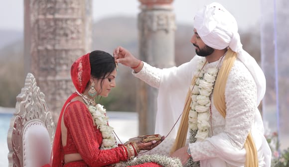 Groom in a white sherwani and turban applying sindoor to the bride, who is seated in a red lehenga on a carved chair, hills behind, at Fateh Vilas, Udaipur.
