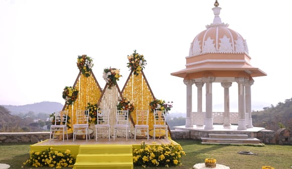 Stage draped with marigold garlands and set with white chairs and wreaths on a lawn beside a domed chhatri pavilion, hills behind, at Fateh Vilas, Udaipur.