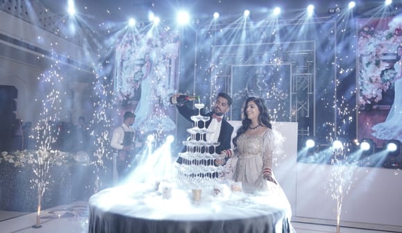 Couple pouring champagne over a tiered glass tower on a draped table, framed by sparkler fountains, stage lighting and floral panels at Fateh Vilas, Udaipur.
