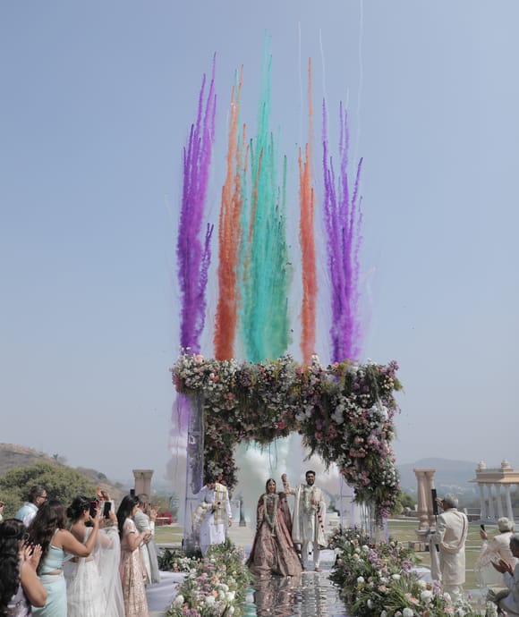 Bride and groom beneath a floral mandap as jets of purple, teal and orange smoke rise behind them, guests lining the walkway at Fateh Vilas, Udaipur.