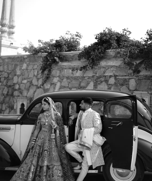 Black and white photograph of a bride in an embroidered lehenga beside a vintage car as the groom steps out, stone wall behind, at Fateh Vilas, Udaipur.