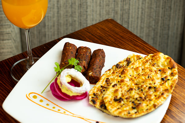 Food: three seekh kebabs with garnish and naan beside a glass of orange drink