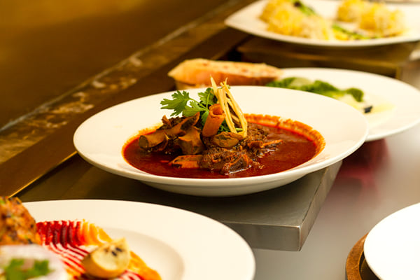 Food: buffet counter with mutton curry bowl and several plated dishes in warm light