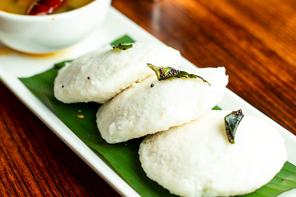 Food: three idlis on banana leaf with a bowl of sambar on a wooden table