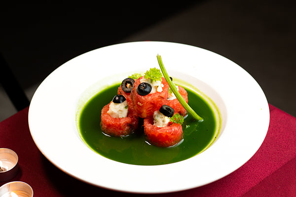 Food: watermelon pieces topped with cheese and olives in green sauce bowl