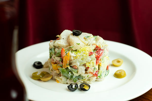 Food: stacked creamy salad with olives, peppers and greens on a plate against maroon cloth