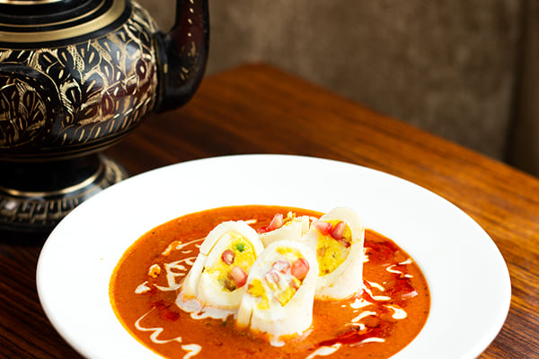 Food: stuffed egg-white rolls in orange curry sauce in a white bowl beside a brass pot