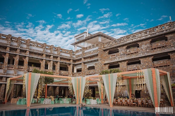 Wedding celebration: draped canopy mandap beside a pool with stone resort building behind