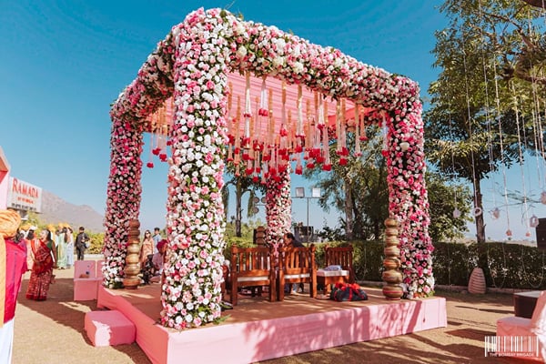 Wedding celebration: pink platform mandap covered in flowers outdoors with guests and trees behind