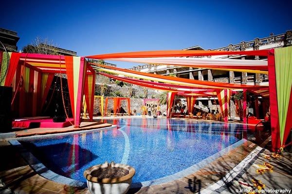 Wedding celebration: pool with orange and red draped canopies and flower bowl under blue sky