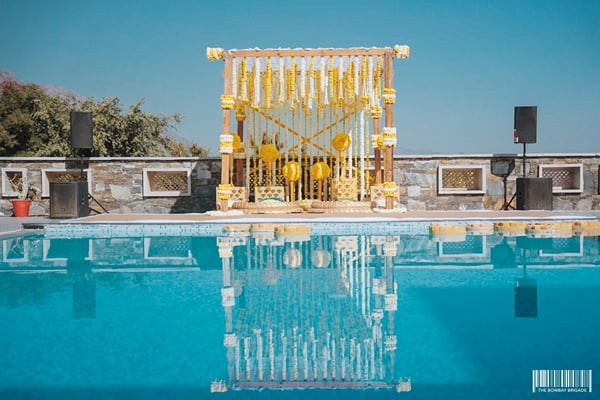 Wedding celebration: yellow flower-curtain mandap beside pool with speakers and stone wall